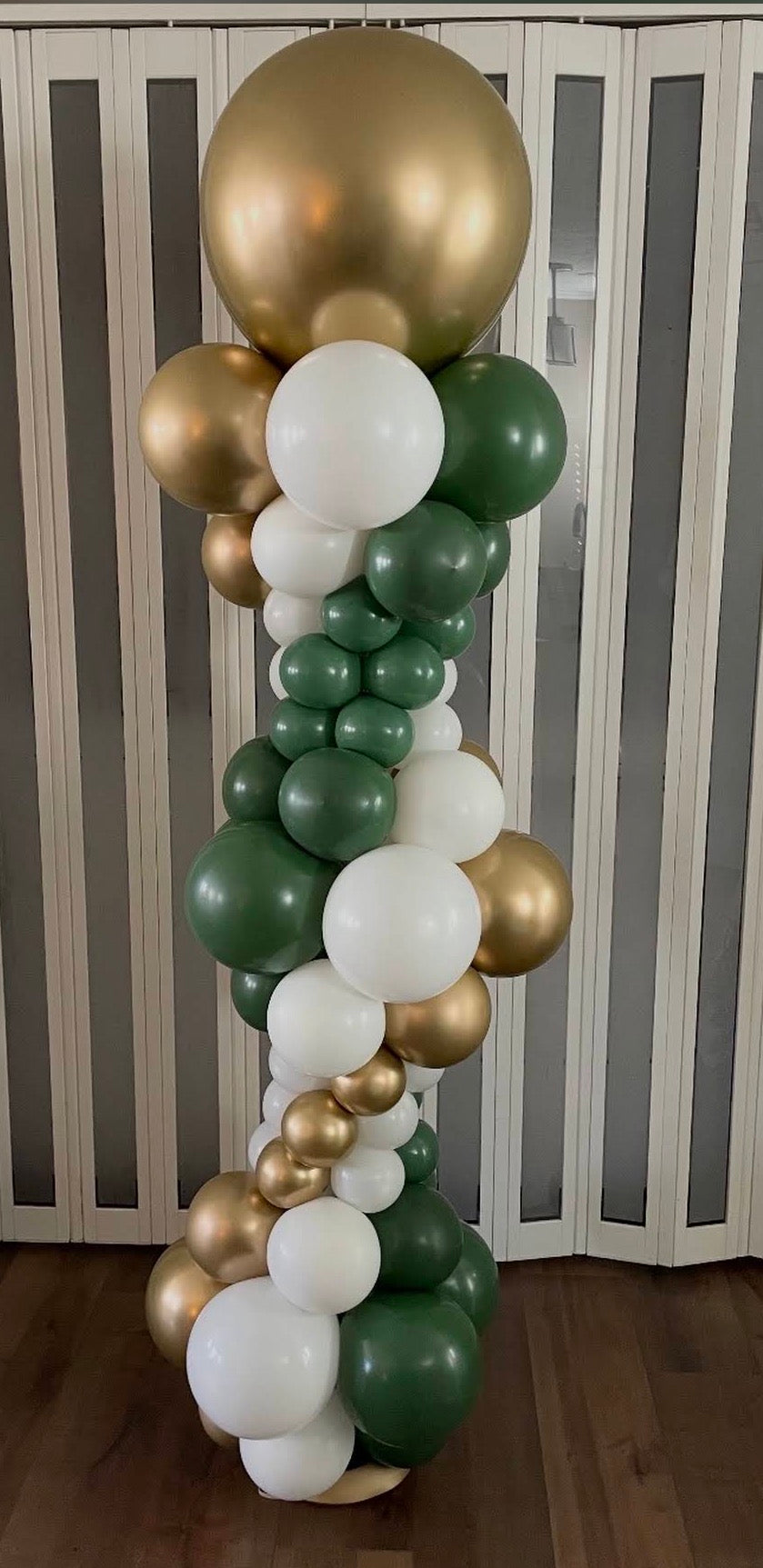 A decorative balloon garland pillar featuring a mix of large, medium, and small matte balloons in metallic gold, deep green, and white, arranged against a white slatted folding screen and dark wood floor.