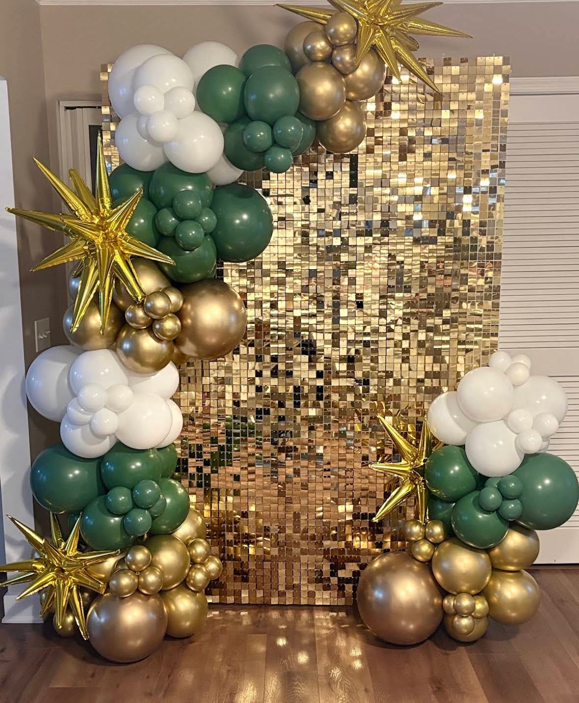 A festive balloon garland arch setup in green, white, and metallic gold, featuring starburst and cloud balloon clusters flanking a large gold sequin backdrop in an indoor room.