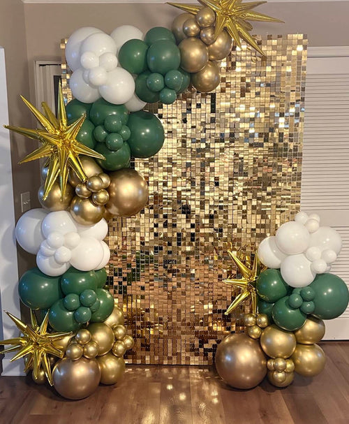 A festive balloon garland arch setup in green, white, and metallic gold, featuring starburst and cloud balloon clusters flanking a large gold sequin backdrop in an indoor room.
