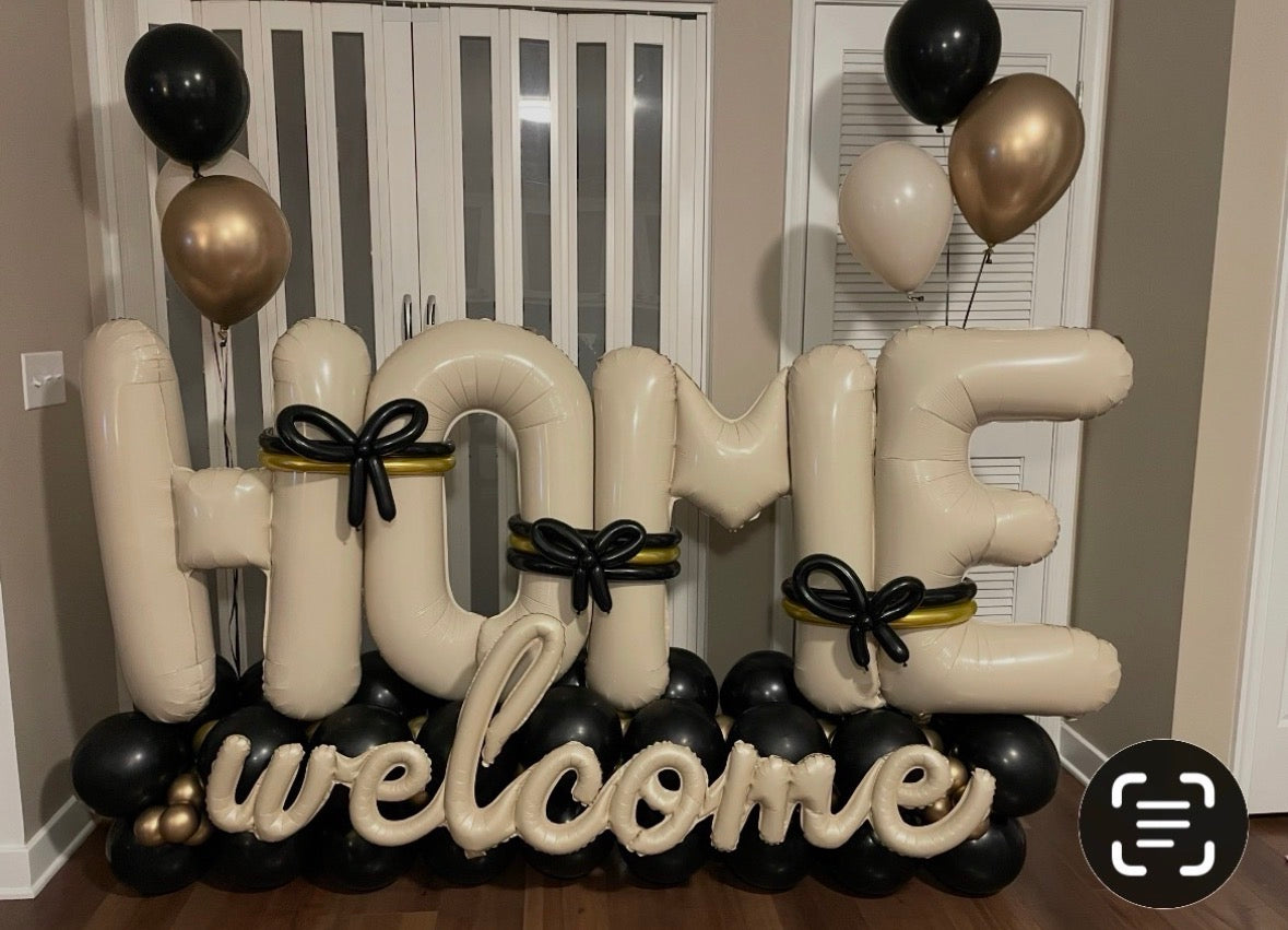 Large inflatable letter balloons spelling 'HOME welcome' in cream, black, and metallic gold, arranged indoors with matching balloon clusters near white louvered doors.