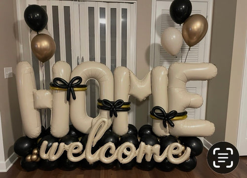 Large inflatable letter balloons spelling 'HOME welcome' in cream, black, and metallic gold, arranged indoors with matching balloon clusters near white louvered doors.